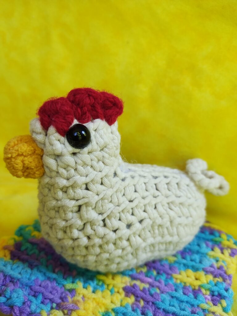 Crocheted cream-colored chicken with a yellow beak and red comb against yellow backgroud